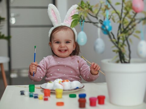 A dark-haired girl 1.5 years old with rabbit ears on her head sits at a white table with a brush and paints paints Easter eggs, next to her on the branches is a green plant on which colored eggs hang.