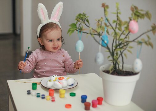 A dark-haired girl 1.5 years old with rabbit ears on her head sits at a white table with a brush and paints paints Easter eggs, next to her on the branches is a green plant on which colored eggs hang.