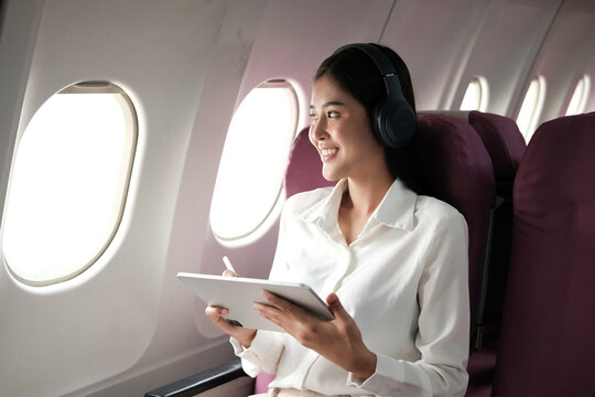 Cheerful Asian woman sitting on a plane looking out the window and use the tablet while traveling