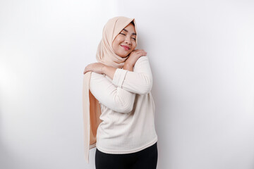 Fototapeta premium Young beautiful Asian Muslim woman wearing a headscarf over white background hugging herself happy and positive, smiling confident. Self-love and self-care