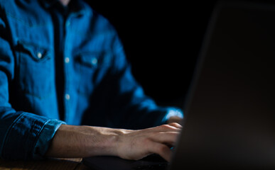 Man working on laptop computer in the dark. Working at night with computer screen light. Blue shirt young business man typing on his computer