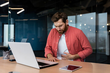 A man in the office at the workplace is sick, he has a severe stomach ache, the businessman got food poisoning from lunch, he is sitting at the table inside the building.