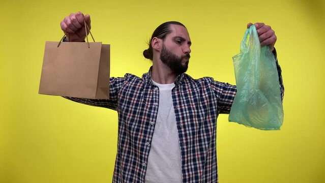 Sensible mid adult man comparing eco-friendly paper bag VS plastic bag, holds arms scales, choosing recyclable reusable packaging isolated on yellow background indoors. Environment protection concept