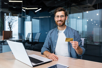 Successful businessman inside office building at workplace smiling and looking at camera, mature adult man holding and showing bank credit card, smiling contentedly.