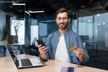 A smiling young man is holding a phone and a credit card. Stands in the office, dials on the phone,...
