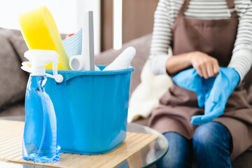 Woman in apron puts on rubber gloves and ready to housework sitting in living room interior. Housewife with cleaning equipment has fun and many household chores, professional cleaning service.