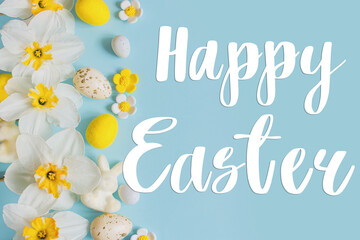 Happy Easter text sign on stylish Easter eggs, bunny and yellow daffodils flowers flat lay on blue background. Modern Easter greeting card. Handwritten lettering