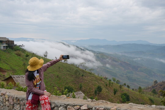 Tourists use their mobile phones to take pictures of fog-shrouded mountain views. Woman wearing purple-brown robe wearing yellow wide-brimmed hat and trousers with Red elephant pattern sit on chair.
