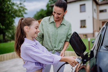 Happy mother showing her little daughter how to charge their electric car.