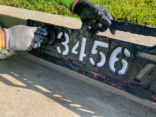 City worker painting address on street curbs using stencil numbers with white and blank paint
