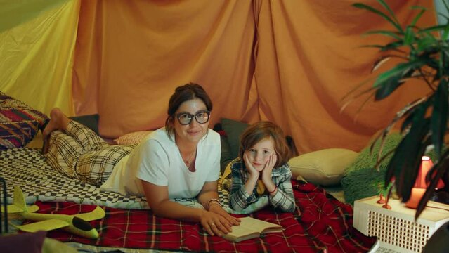 A kind hearted mother wearing glasses and her adorable son are in a huge blanket fortress and they are laying down staring at the camera directly and smiling