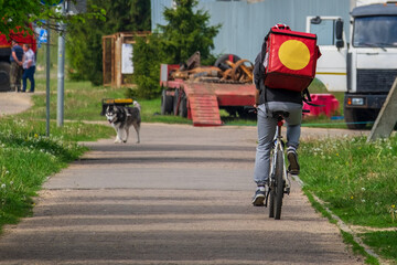 Naklejka premium Food delivery to customers at home. A delivery service worker with a thermal backpack on a bicycle quickly delivers food from a restaurant, supermarket or cafe. Online food ordering
