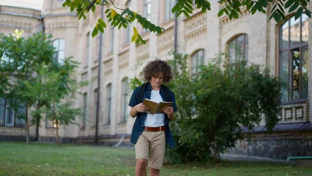 Satisfied Caucasian schoolboy walking on school backyard reading book smiling. Portrait of happy boy with backpack enjoying education leaving in slow motion. Knowledge and confidence concept
