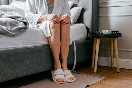 Close Up Of Lonely Elderly Woman Sitting On The Bed Posing With Legs Holding Knees In Arms In Front Of Camera At Home Against Bedroom.