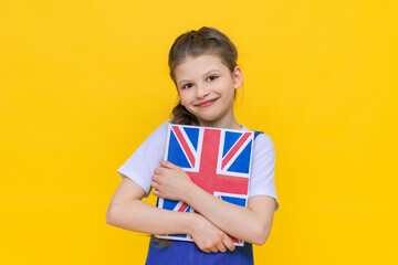 Learning English for children. A little girl is holding a book with an English flag in her hands and smiling broadly, on a yellow isolated background.