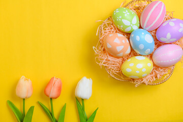 Happy Easter holiday greeting card design concept. Colorful Easter Eggs and spring flowers on yellow background. Flat lay, top view, copy space.