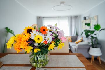 Kitchen counter table with focus on vase with huge multicolor various flower bouquet with blurred background of modern cozy living room with couch and green plants. Open space home interior design.