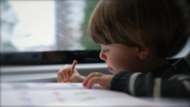 One focused child doing school activity seated in a moving train by window. Little boy nose picking while studying on commute