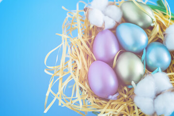 Fototapeta na wymiar Colored Easter eggs in a nest on a bright blue festive background. Sprigs of cotton lie next to a wicker basket. Top view, place for text. Easter concept.