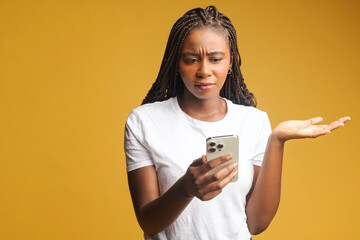 Serious pensive woman of young age holding smartphone in hands, dont understand, chatting with friends or making food order, has concentrated facial expression isolated on yellow background