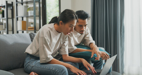 Happy young Asian couple sit on couch fun argument use laptop online shopping at home on holiday....