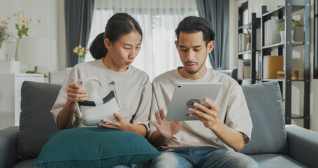 Young Asian couple sit on couch hold digital tablet and wear virtual reality glasses are watching and show imagine spend time together have fun at home on weekend. Lifestyle tech innovation concept.