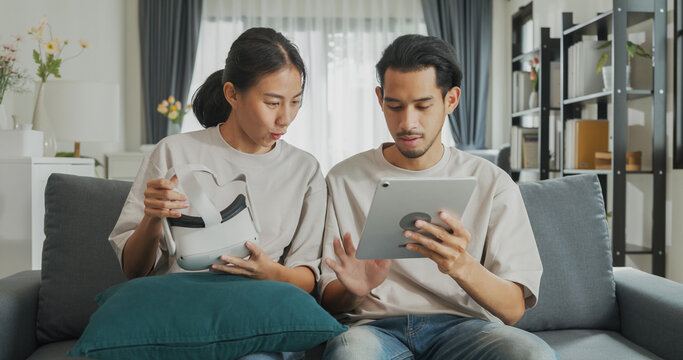 Young Asian couple sit on couch hold digital tablet and wear virtual reality glasses are watching and show imagine spend time together have fun at home on weekend. Lifestyle tech innovation concept.