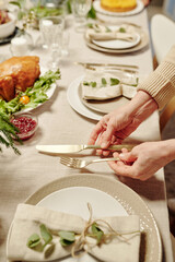 Hands of mature woman putting clean fork and knife on served table with plates, napkins, wineglasses, homemade food and decorations