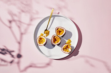 Fresh passion fruit on white ceramic plate with golden spoon. Sun light, shadows, top view. Light pink background. Summer exotic breakfast.