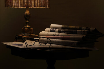 an old lamp illuminates a table on which there are newspapers and spectacles