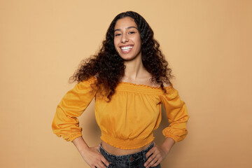 Happy smiling confident latin student girl dressed to go on summer party, posing with hands on waist against brown studio background, feeling proud and satisfied with her fashionable look