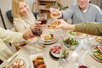 High angle of hands of intercultural family members toasting with glasses of homemade wine over table served with appetizing food