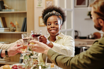 Happy young African American woman looking at her husband while clinking with wineglasses of family members during toast