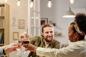 Happy young man looking at his African American wife with smile while clinking with wineglasses of family members during celebration