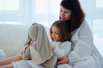 Portrait of happy family. Young mother and little daughter sits in pajamas on a couch with a soft toy at home.