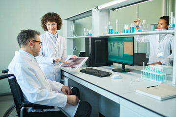 Confident female researcher showing male colleague with disability macro image of virus cell during discussion of new variant in laboratory