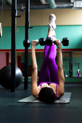 Fototapeta na wymiar Young woman doing exercises with dumbbells on a mat in the gym. Beautiful brunette in a purple suit. Activity, sports and beauty. Vertical.