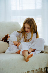Portrait of the adorable little girl in white old-fashioned pajama playing with white doll on the sofa at home