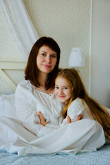 Young mother and little cute daughter sits on the bed in the bedroom in old fashioned white pajamas in the morning