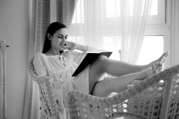 Black and white image of a young brunette woman is relaxing at home in the morning. She reads a book while in daylight near the window.