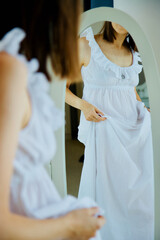 A young adult woman in white pajama in front of mirror. She and looks at herself in the reflection, cropped rear view.