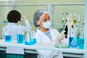Fototapeta premium Young female virologist or pharmacologist holding flask with blue liquid chemical substance during scientific experiment in laboratory