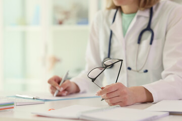 Doctor writes notes holding glasses at table in hospital