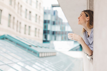 Fototapeta premium Young woman looking out the window and holding mug at home or hotel room. She drinking coffee or tea after wake up in the morning. Good morning! Woman look happy, positive mood, sitting on windowsill.