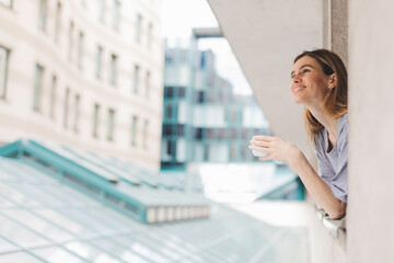 Fototapeta premium Young woman looking out the window and holding mug at home or hotel room. She drinking coffee or tea after wake up in the morning. Good morning! Woman look happy, positive mood, sitting on windowsill.