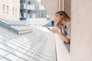 Young woman looking out the window and holding smart phone at home or hotel room. Woman using mobile phone chatting, scrolling, read news in the morning. Woman look puzzled and doubtful, thoughtful.