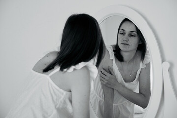 Portrait of young adult woman in white pajama in front of mirror. She and looks at herself in the reflection, rear view. Black and white image.