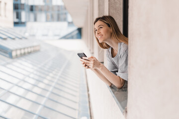 Fototapeta premium Young woman looking out the window and holding mug at home or hotel room. She drinking coffee or tea after wake up in the morning. Good morning! Woman look happy, positive mood, sitting on windowsill.