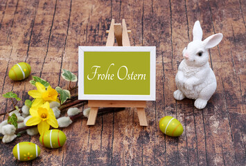 Osterkarte Frohe Ostern. Narzissen und Zweige mit Osterhasen und Eiern . Auf einer Staffelei steht der Text Frohe Ostern.