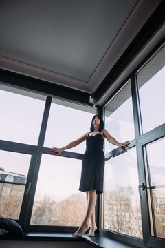 A cute graceful brunette in a light black dress on the background of a panoramic window 4560.